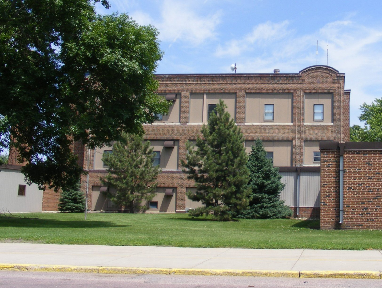 A multi-story brick building with small windows, surrounded by green trees and grass, under a partly cloudy blue sky.