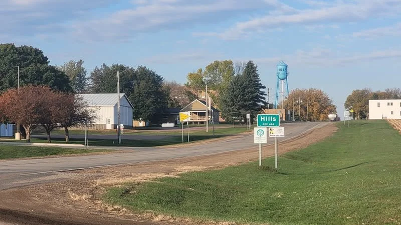A rural street scene with trees, houses, a water tower, and street signs in a small town.