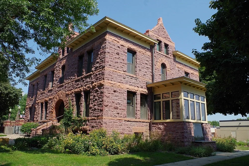 A large, historic stone house with a red sandstone exterior, multiple windows, a small staircase, and a landscaped garden in front. The house has a distinctive turret and an enclosed porch with large windows.
