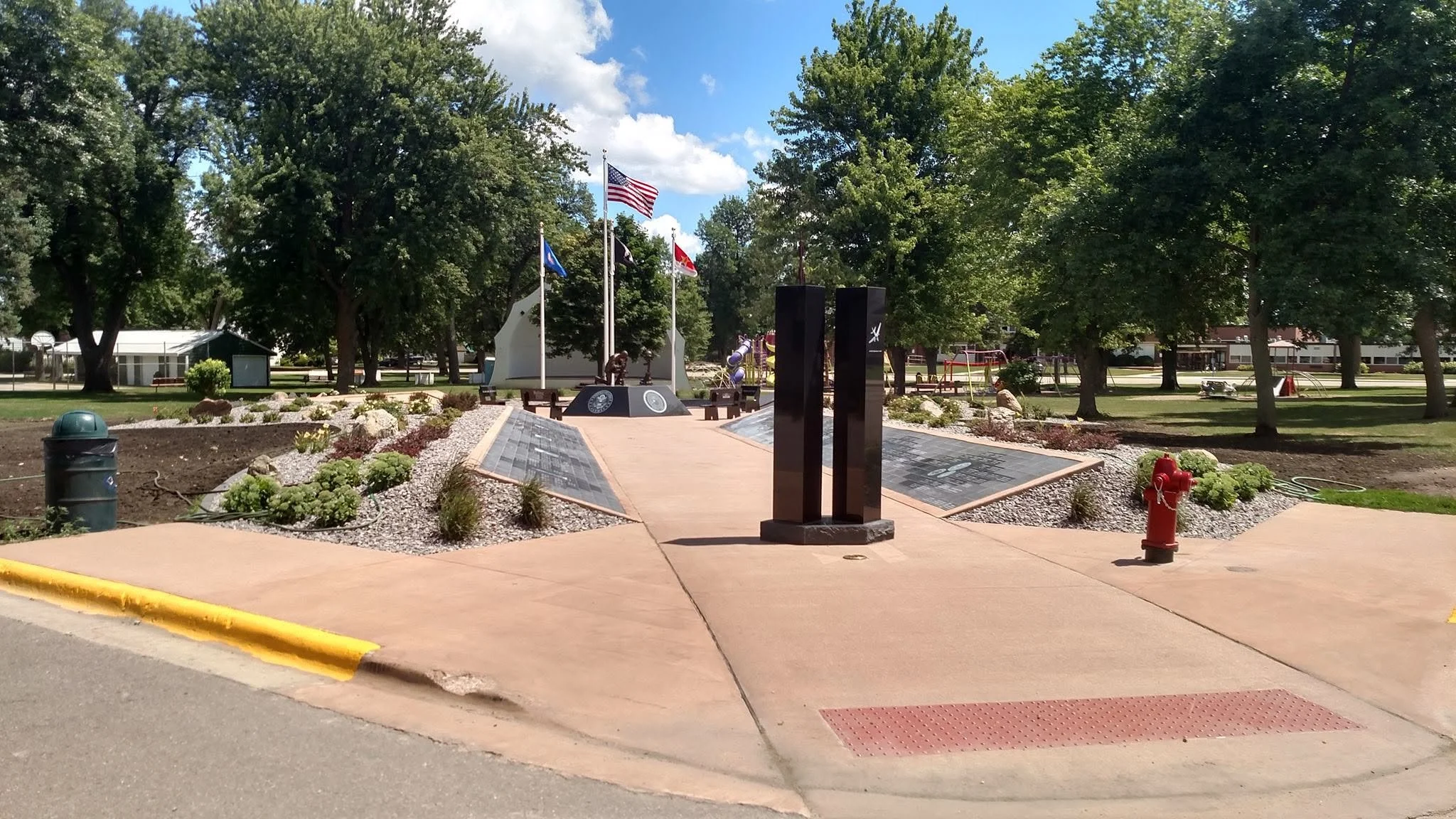 Memorial park with flags, plaques, trees, and a playground in the background under a blue sky with clouds.