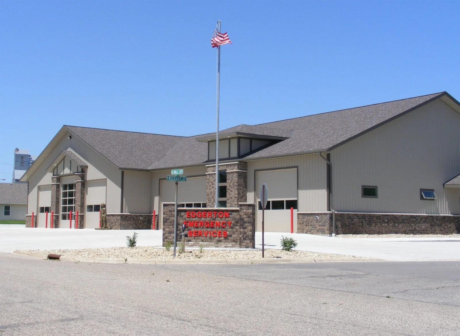 A building labeled Edgerton Emergency Services with American flag on a tall pole in front. The building has beige siding with brick accents and a gabled roof, Located at the corner of E. Mill Street and Mechanic Street.