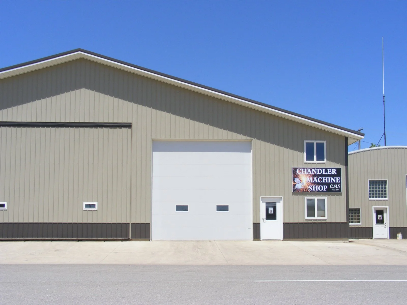 A beige industrial building with a sign that reads 'Chandler Machine Shop' in white letters on a black background. The building has a large white garage door, several small windows, and a door with a sign.