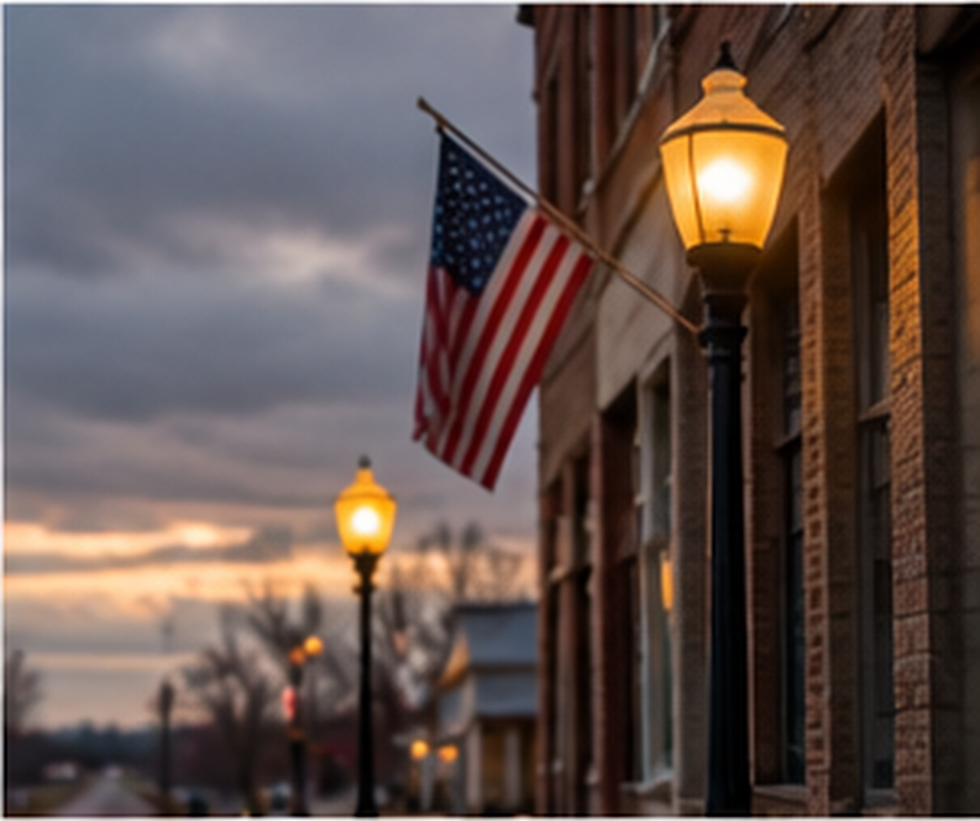 The image depicts a street scene during dusk with a brick building featuring warm illuminated street lamps and an American flag hanging from a pole. The sky is cloudy, and there are silhouettes of trees and additional street lamps in the background.