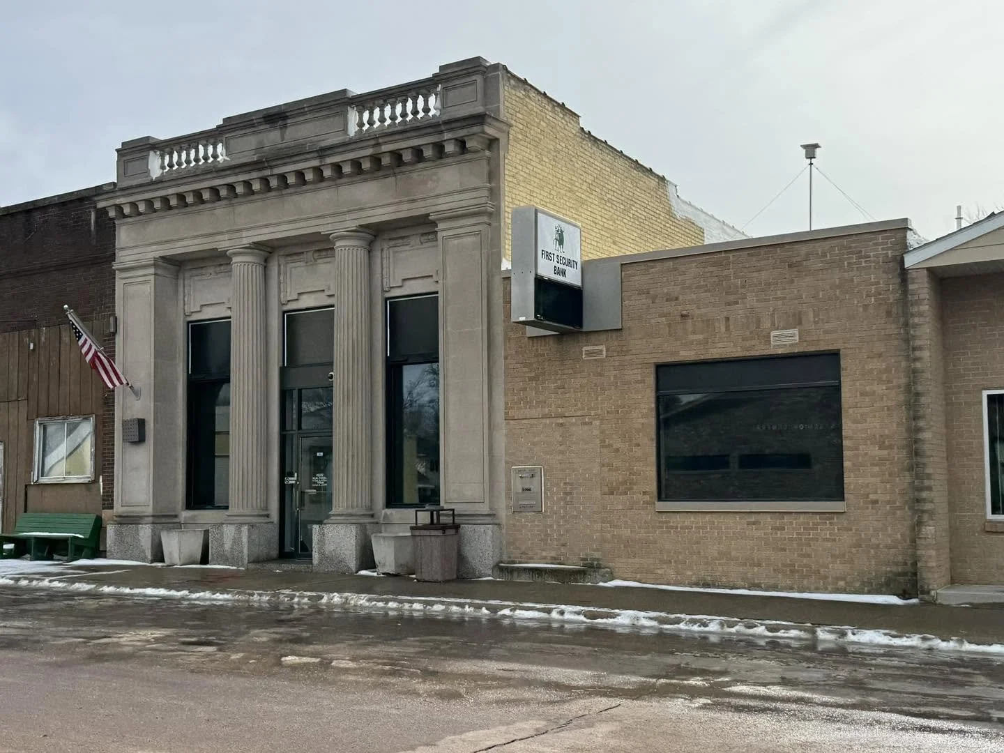 A street view of a small bank building with classic architectural details, including columns and a decorative façade, alongside a modern brick building with a bank sign that reads 'First Security Bank' and an American flag on a front yard.