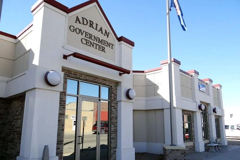 Exterior of the Adrian Government Center building with a sign, glass doors, and a flagpole with a flag.