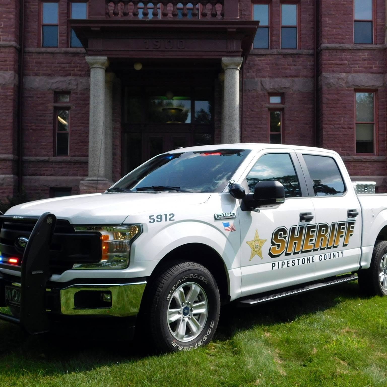 A white sheriff's pickup truck with black lettering and a sheriff's badge parked on a grassy area in front of a red brick building.