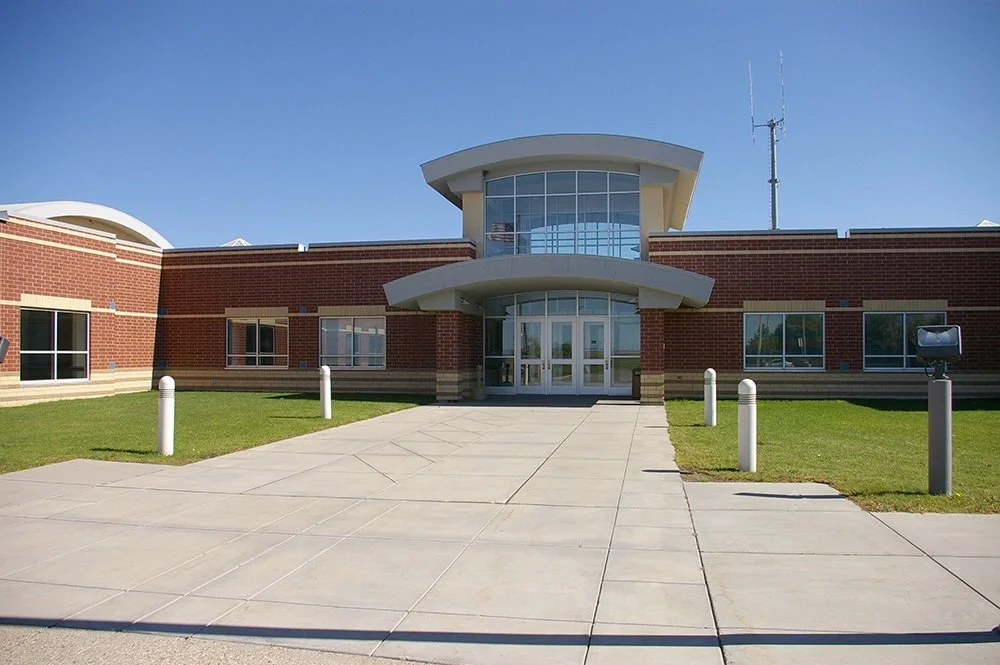The exterior of a modern brick building with a curved entrance and large glass windows, surrounded by a concrete walkway and grass with white posts.