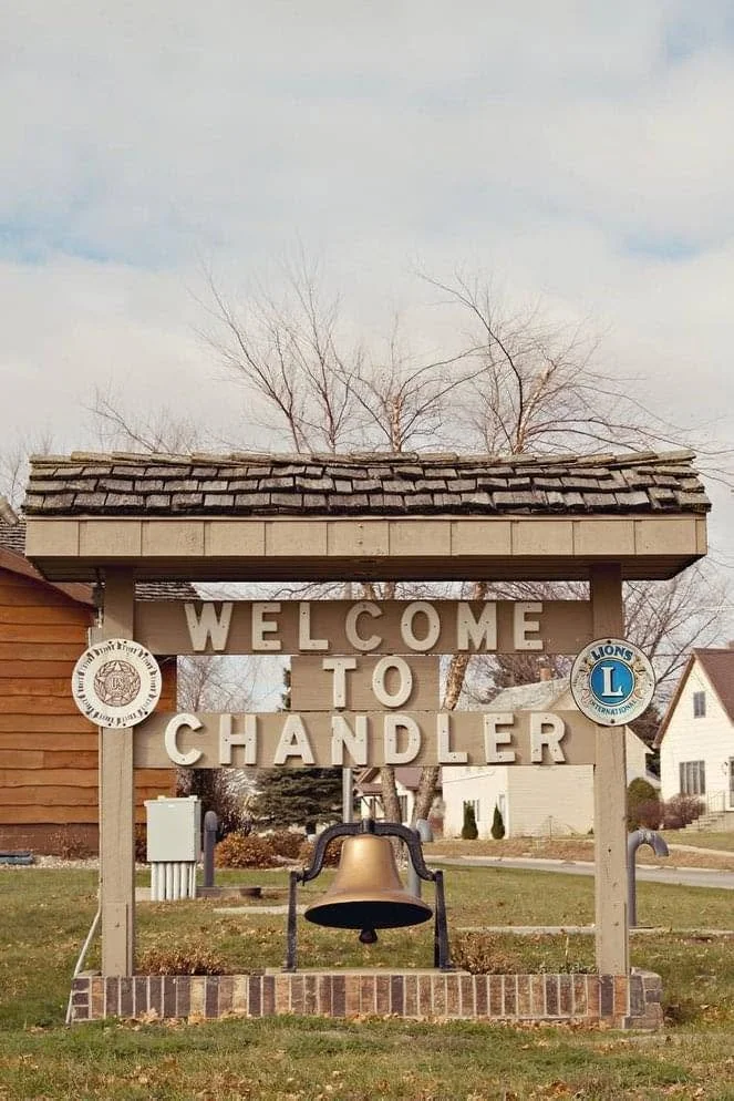 A sign that reads 'Welcome to Chandler' with a large bell hanging below the text, located in a small town setting with houses and leafless trees in the background.