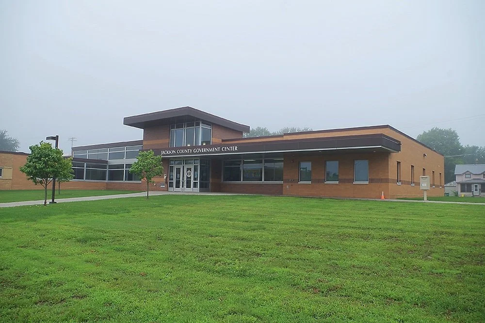 Jackson County Government Center building with glass entrance, brick exterior, and surrounding grass and trees, on a foggy day.