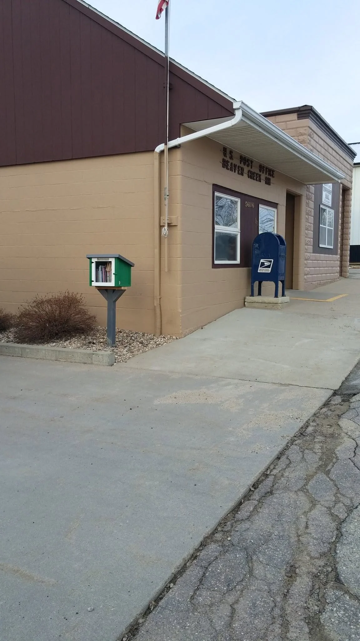 The image shows the exterior of a small post office building with a blue USPS mailbox outside. There is a green public mailbox and a small bush nearby. The building has beige and brown siding with a sign above the window that reads, 'U.S. POST OFFICE BEAVER CREEK MO.'