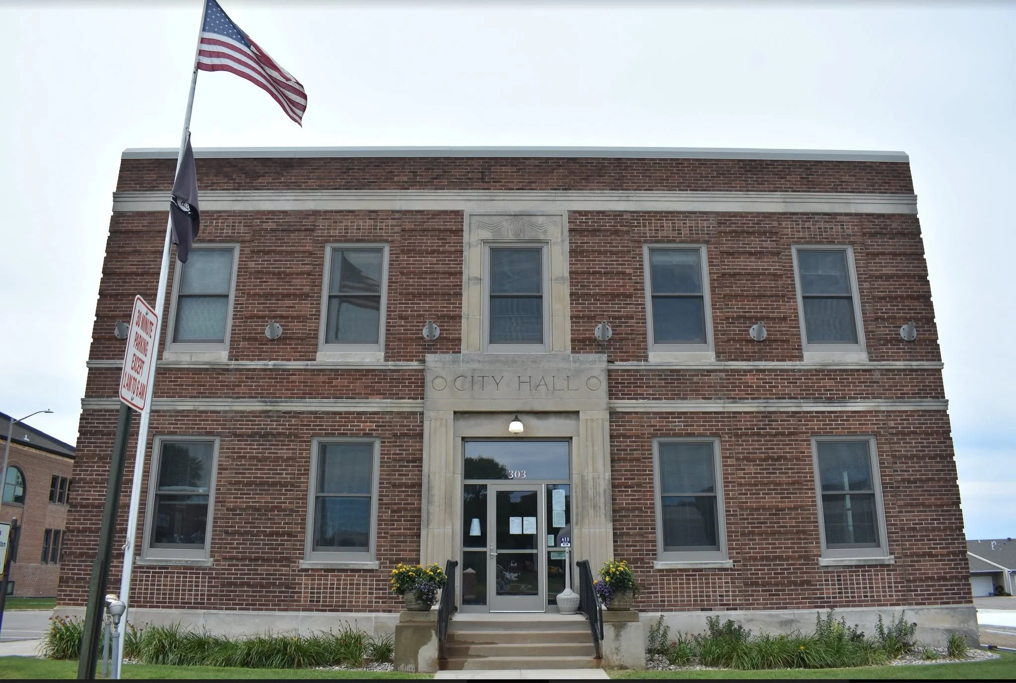Front view of a two-story red brick city hall building with multiple windows, an entrance with stairs, and a sign that reads 'CITY HALL'. An American flag and another flag are flying on flagpoles outside.