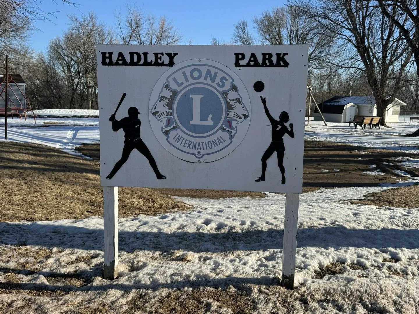 Sign for Hadley Park with Lions International logo and silhouettes of a baseball player swinging a bat and a female volleyball player serving, on a snowy ground with trees and small buildings in the background.