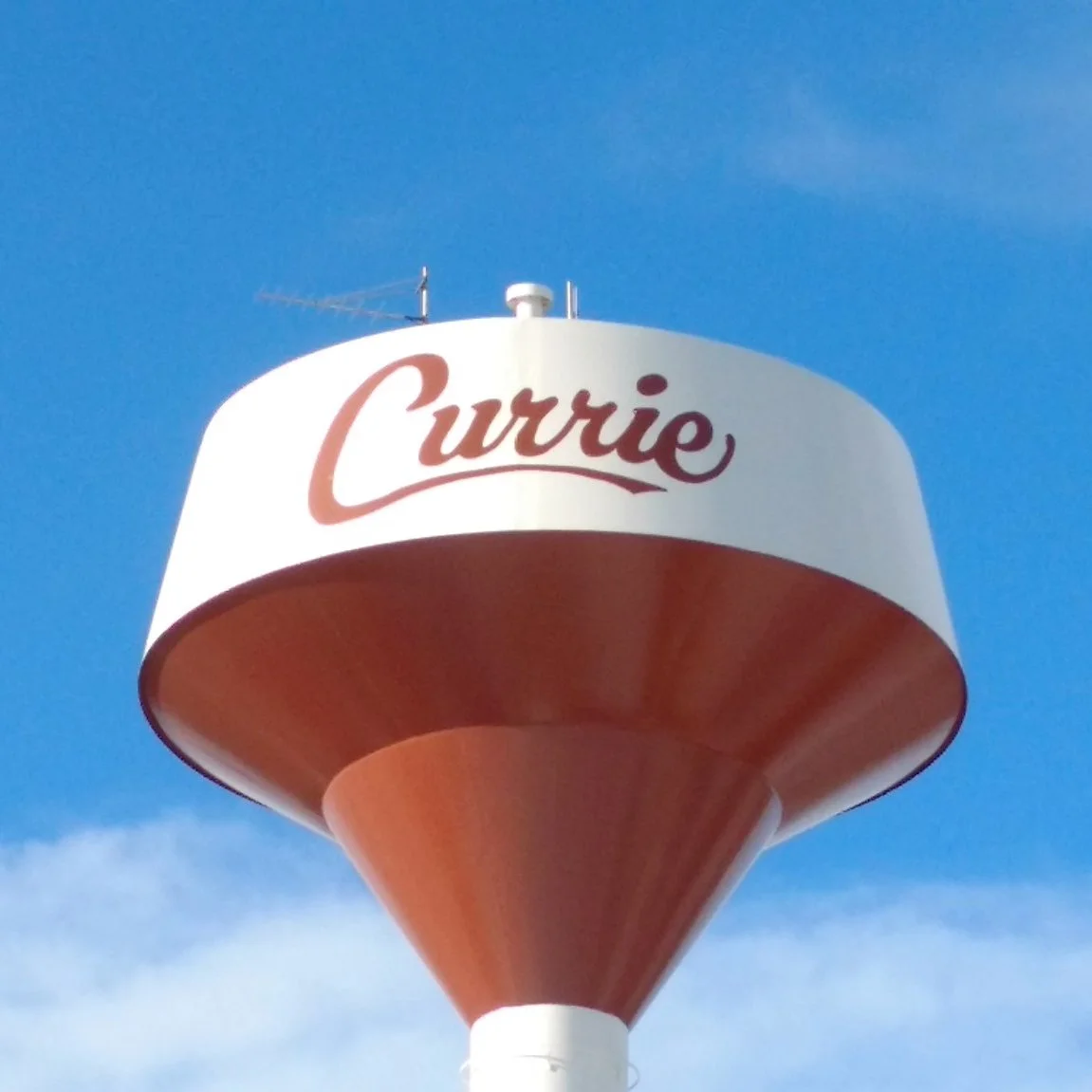 Water tower with the name "Currie" painted in red on the white tank, against a blue sky.