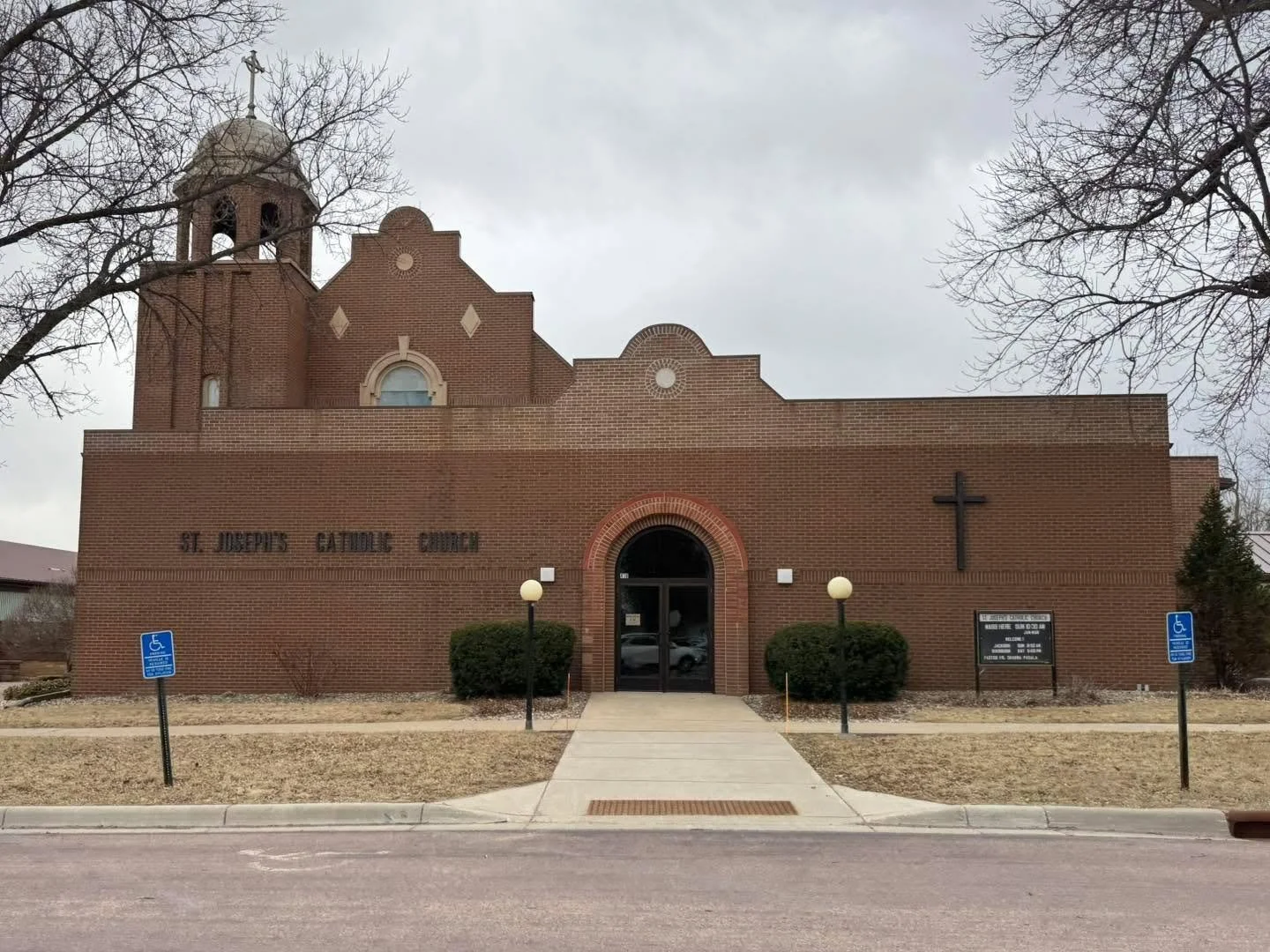 Front view of St. Joseph's Catholic Church, a brick building with a cross on the right side, two wheelchair-accessible signs, two outdoor lamps, and leafless trees.