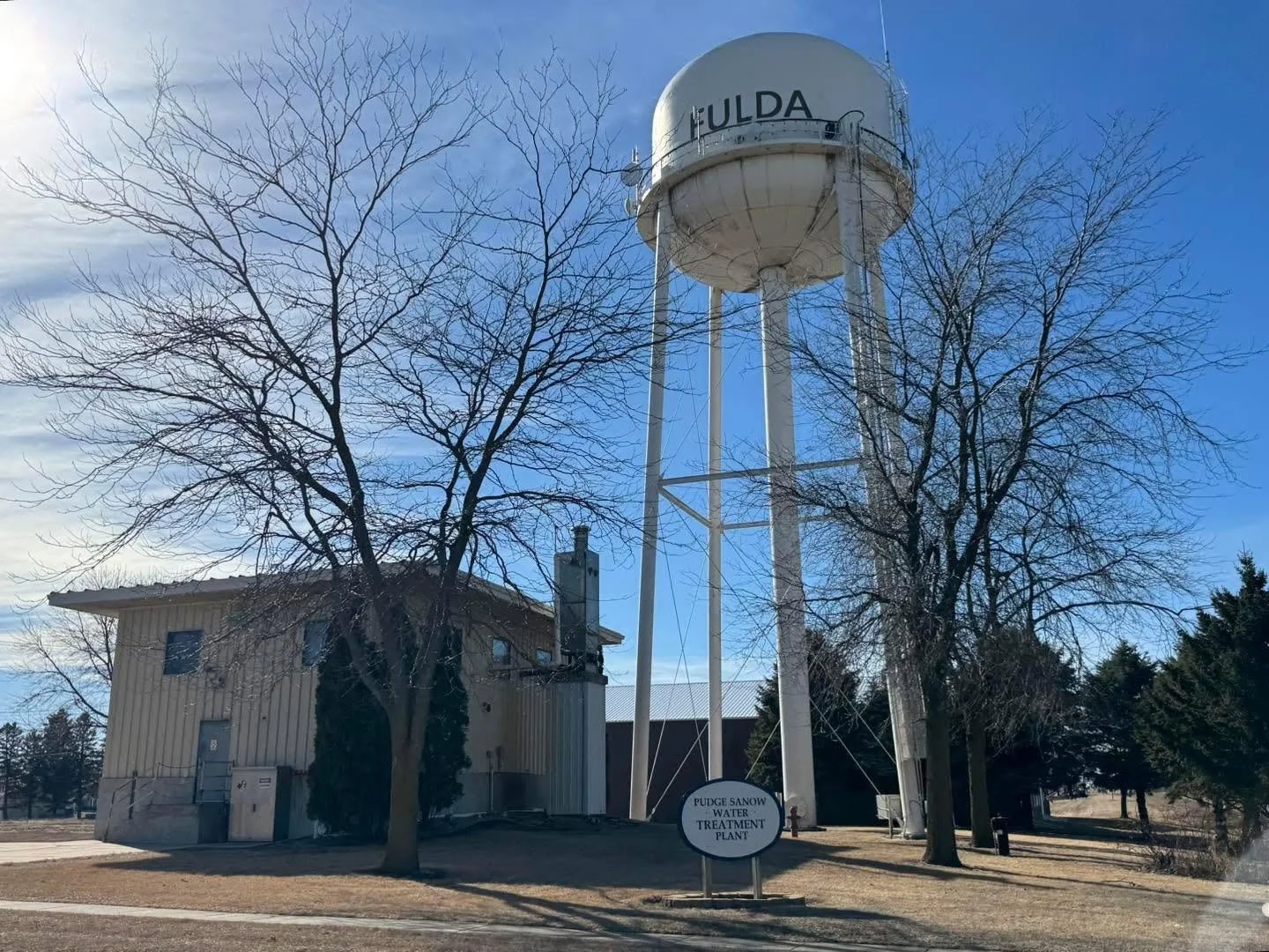 Water tower with the word 'FULDA' on it, next to a small building, trees with no leaves, and a sign that reads 'PUDGE SANOW WATER TREATMENT PLANT,' under a partly cloudy blue sky.