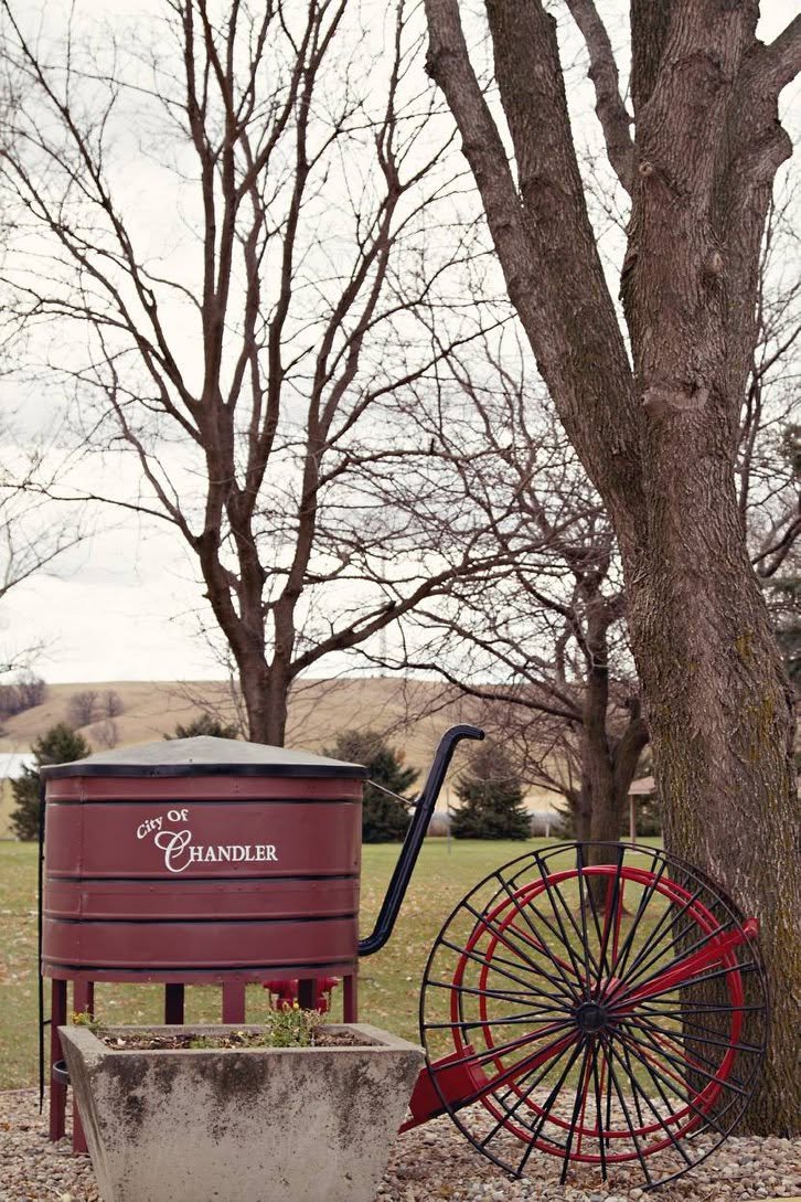 A red water tower with 'City of Chandler' written on it, a decorative red and black wheel, a tree, and a grassy field with hills in the background.