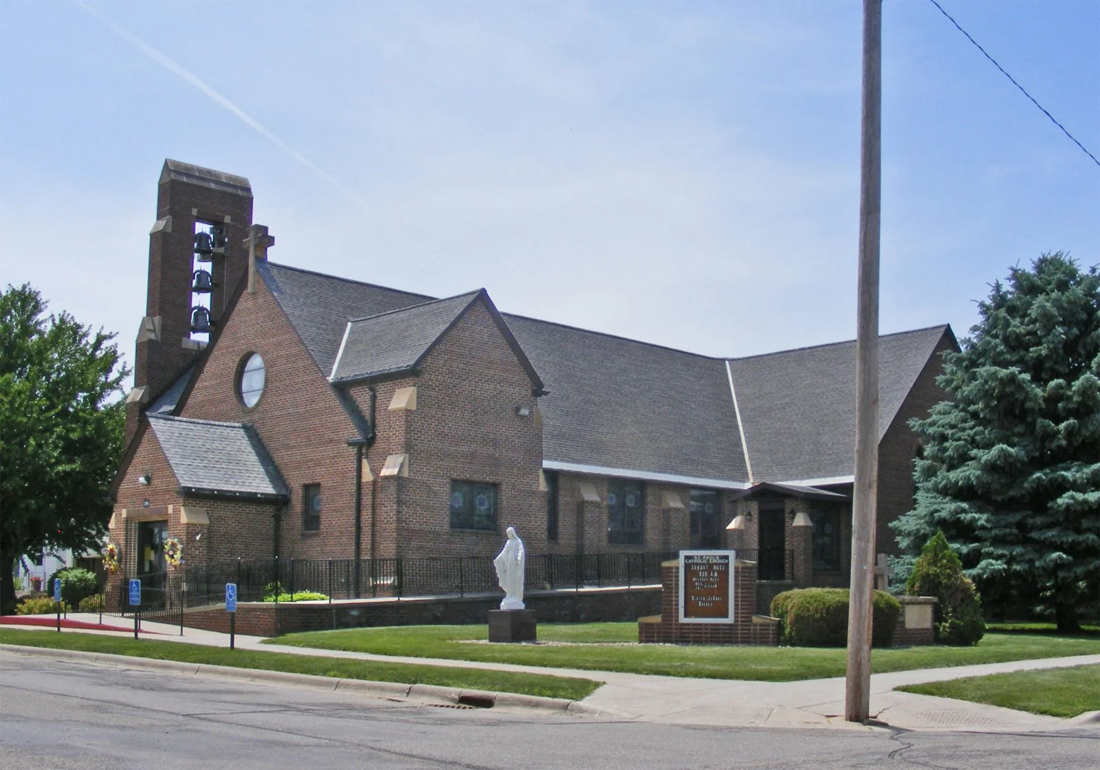A brick church with a tower and bells, a statue of a saint outside, and a sign in front reading 'Catholic Church, 17133 US 175, Temple, Texas'.