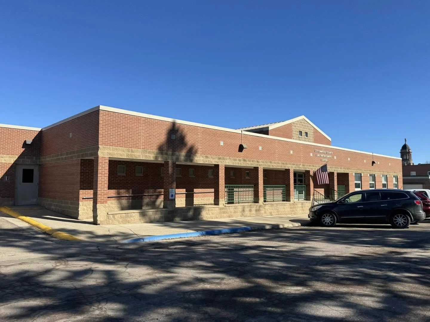 Front view of a red brick community center with a sign that reads "Community Center, Lake Street, Center," with American flag near the entrance, parked cars, and a clear blue sky.