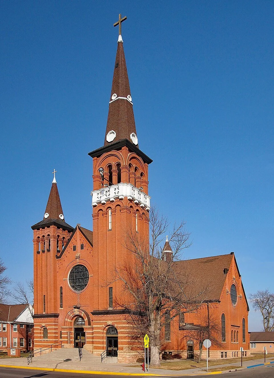Red brick church with tall steeple topped with a cross, leafless tree in front, and clear blue sky.
