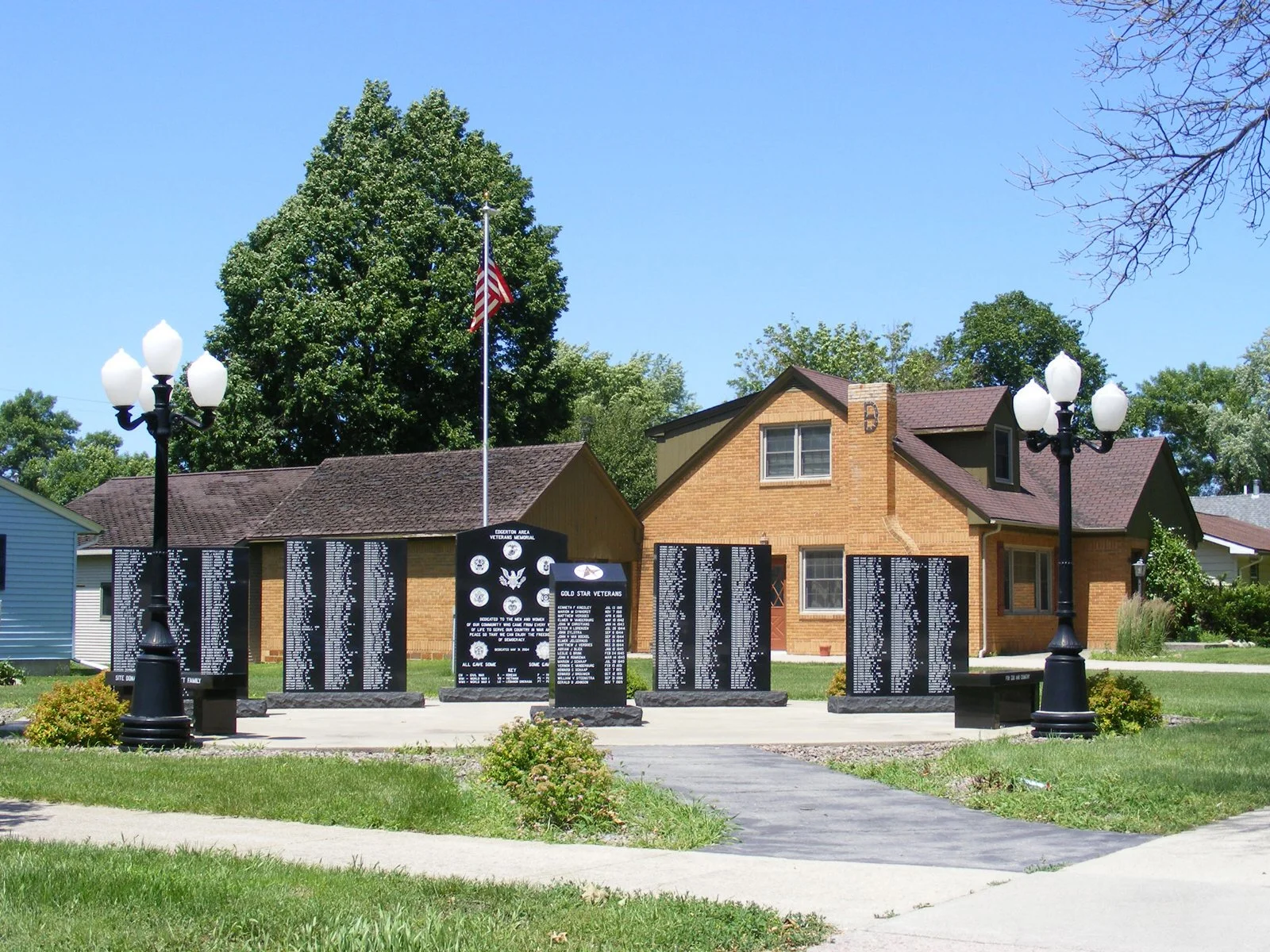 Memorial honoring veterans with multiple black plaques with white text, American flag, two black lampposts with white globes, green grass, trees, and a brick house in the background.
