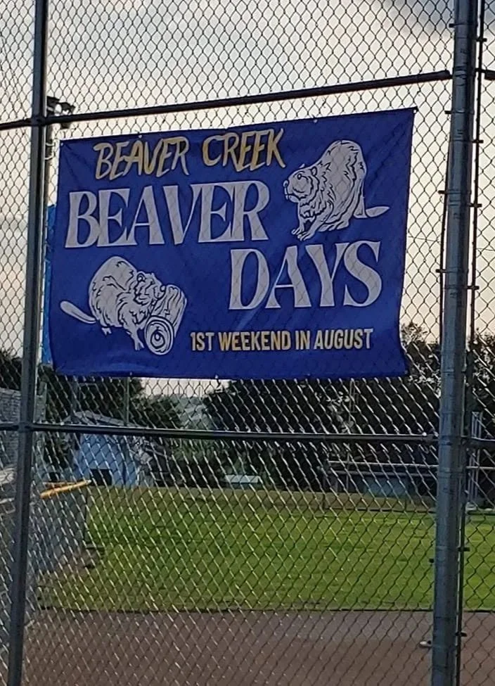 A banner hanging on a chain-link fence advertising the 'Beaver Creek Beaver Days,' which take place on the first weekend in August. The banner features illustrations of a beaver and a bearded dragon.