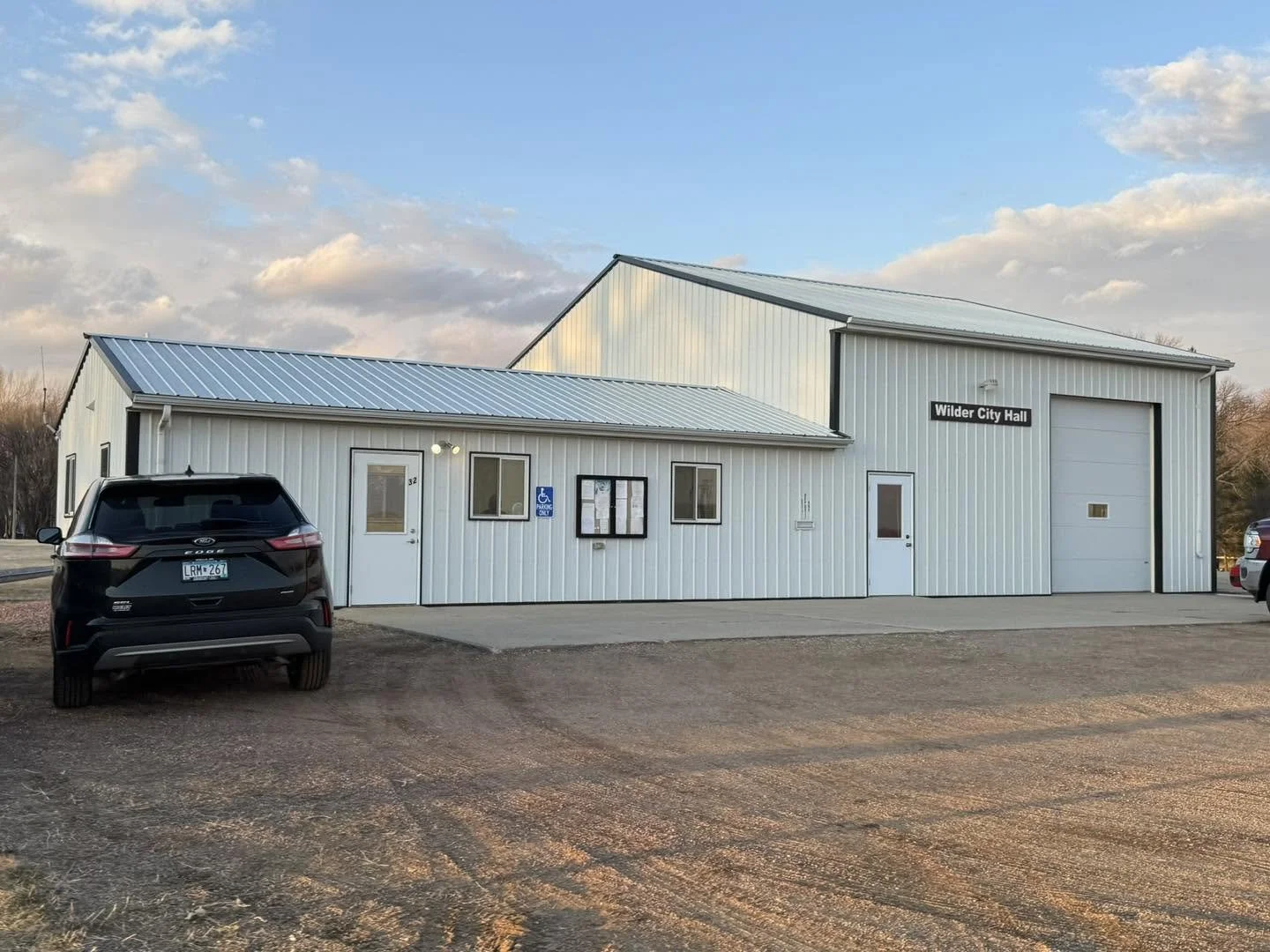White metal building with a sign that says Wilder City Hall, a black SUV parked in front, and a gravel parking area under a partly cloudy sky.