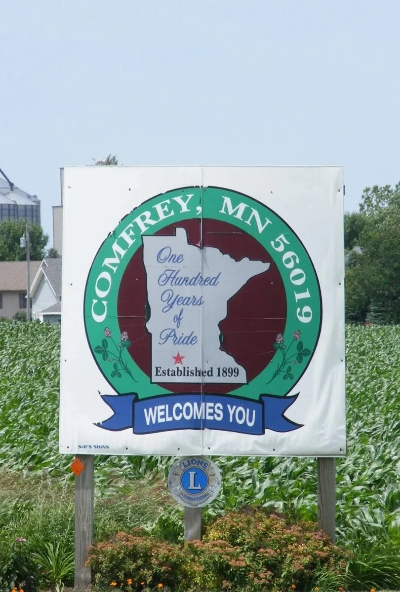 Sign welcoming visitors to Comfrey, Minnesota, celebrating 100 years of pride since 1899, with a map of Minnesota and decorative plants in the foreground.