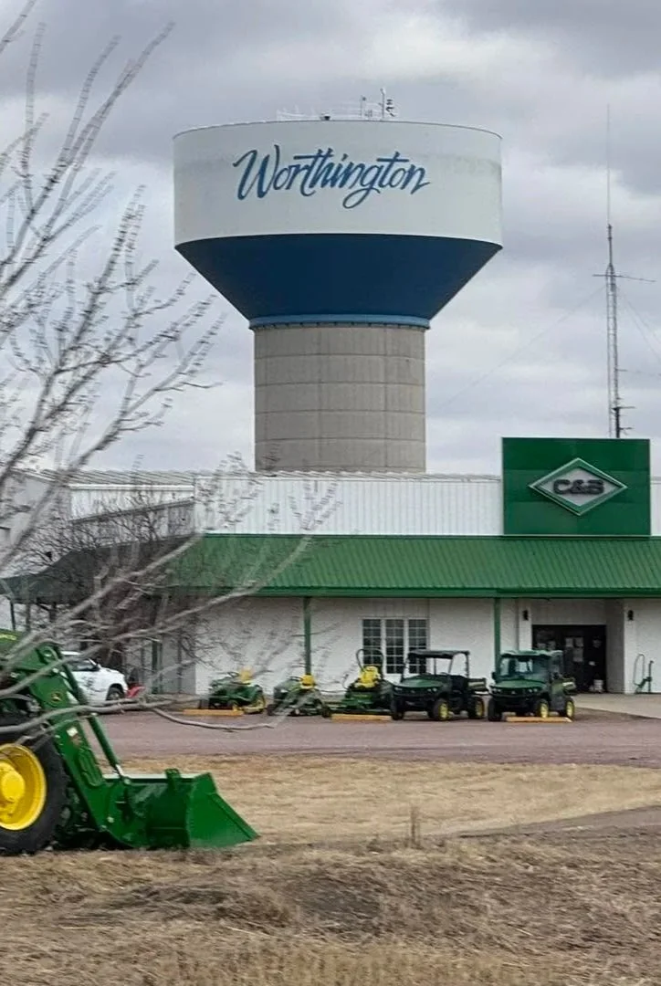 A water tower in Worthington, Ohio, with the town name written on it. In the foreground, there are green tractors and farm equipment parked near a building that has a green roof and a sign with the logo 'C&S'.