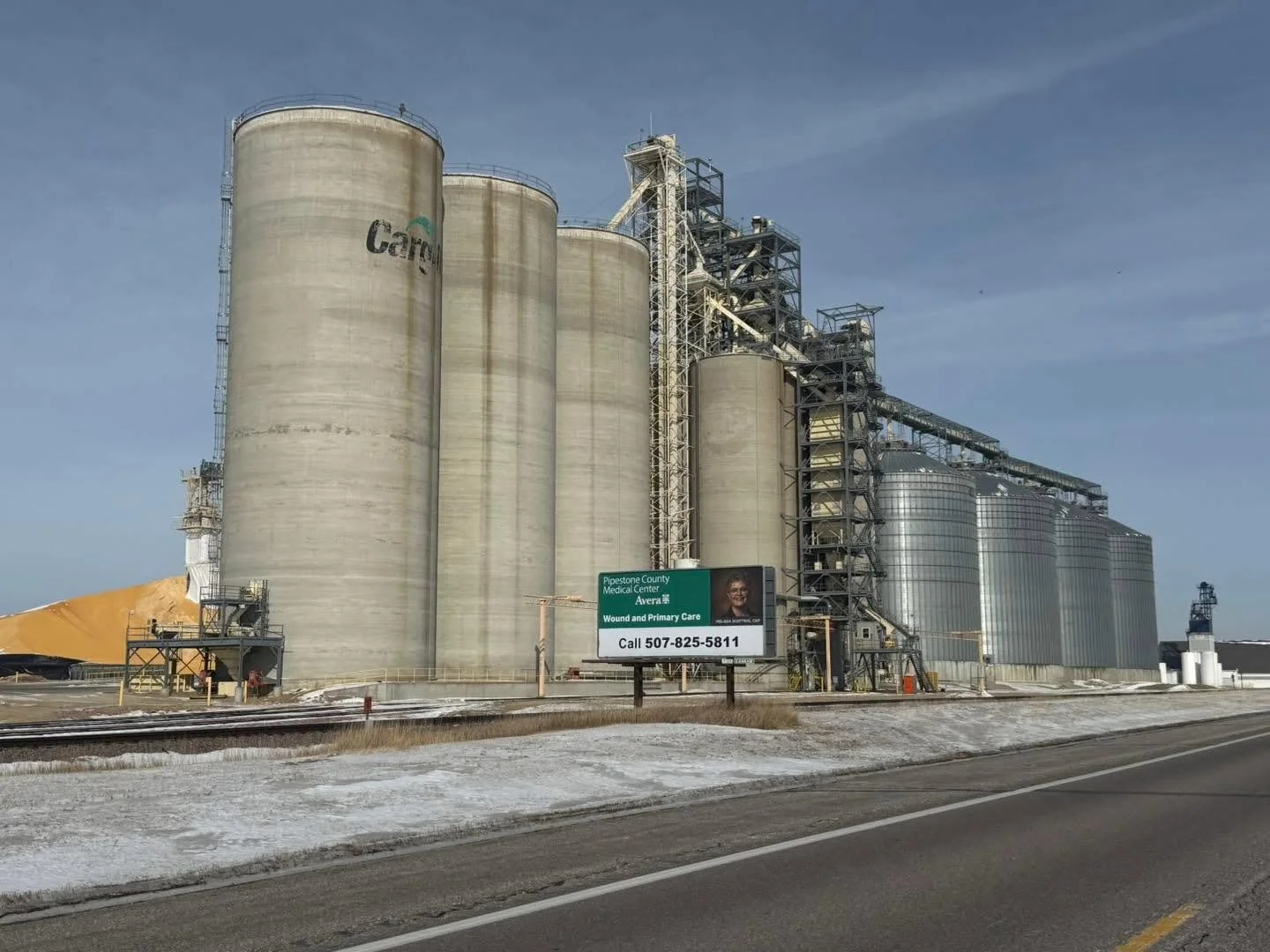 Industrial grain storage silos next to a road with a billboard for Pipestone County Medical Center, featuring a woman and contact information.