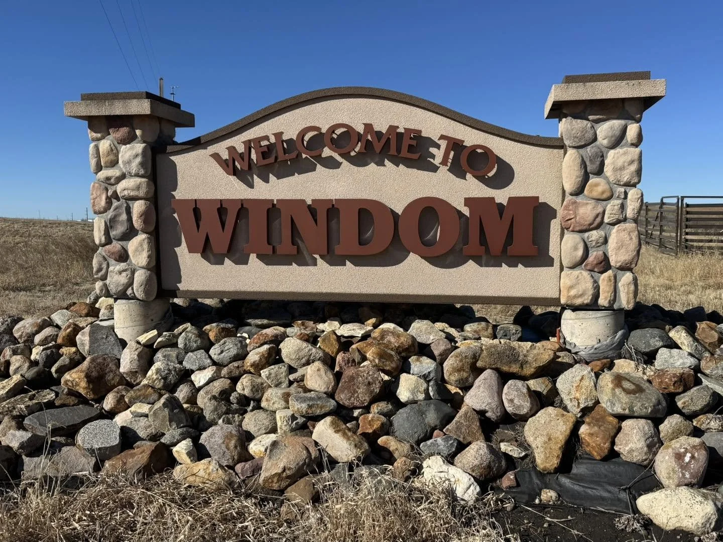 A stone and stucco welcome sign reading 'Welcome to Windom' with a clear blue sky in the background.