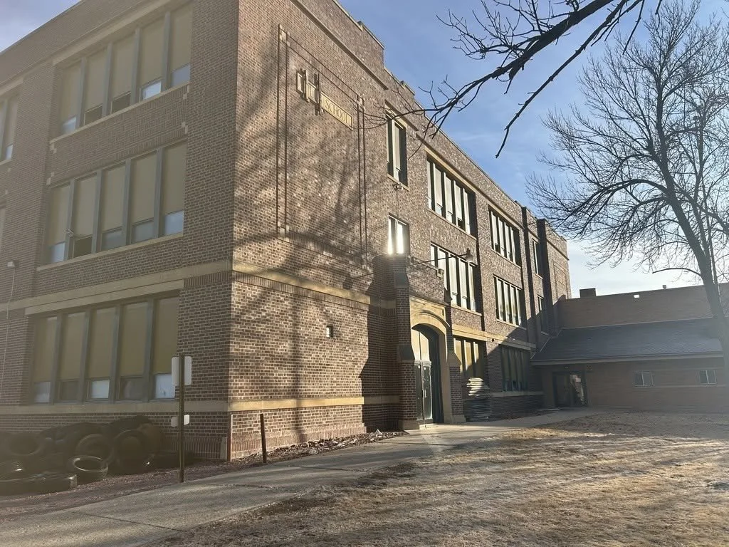 A multi-story brick building with large windows and a small sidewalk in front, with leafless trees casting shadows on the building, under a blue sky with some clouds.