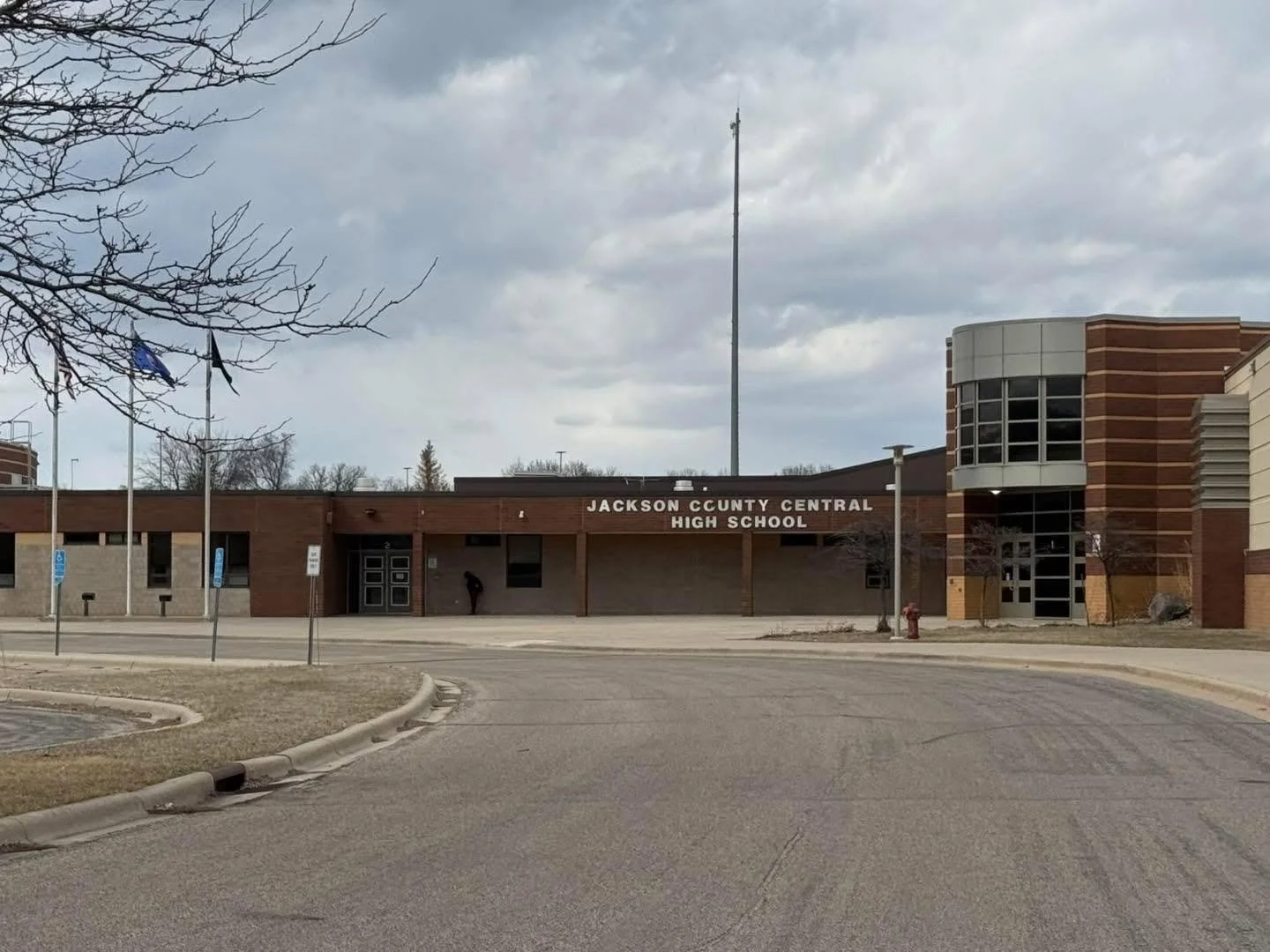Exterior view of Jackson County Central High School building with a brick facade, flags, and a parking lot in the foreground under a cloudy sky.