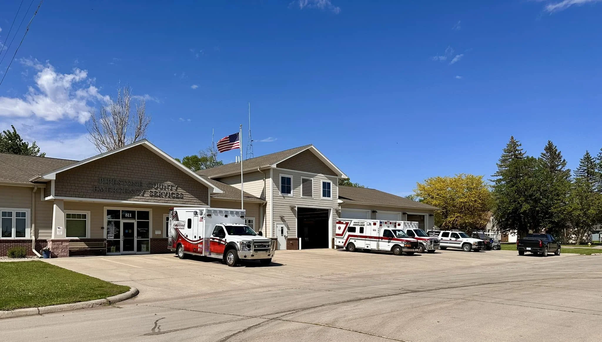 Pipestone County Emergency Services building with emergency vehicles parked in front, American flag on pole, clear blue sky, trees with some foliage.