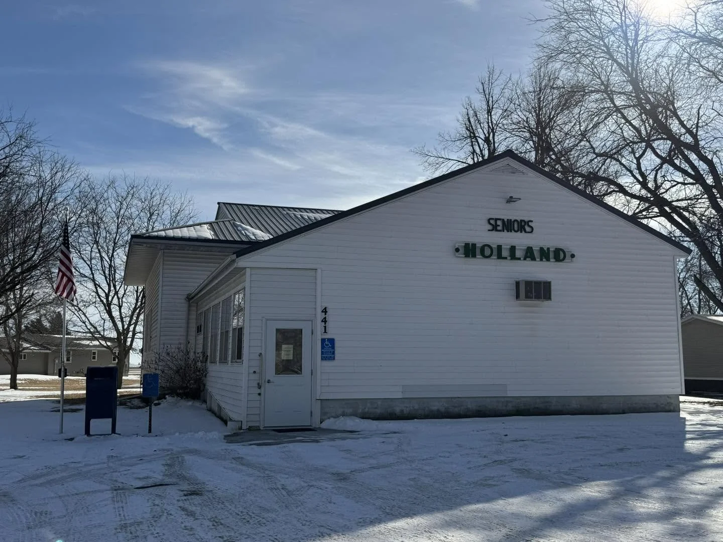 White building with a sign that reads 'Seniors Hollanda' and the street number 441. There is a blue handicap parking sign and a flagpole with an American flag. Snow covers the ground, and leafless trees are in the background under a partly cloudy sky.
