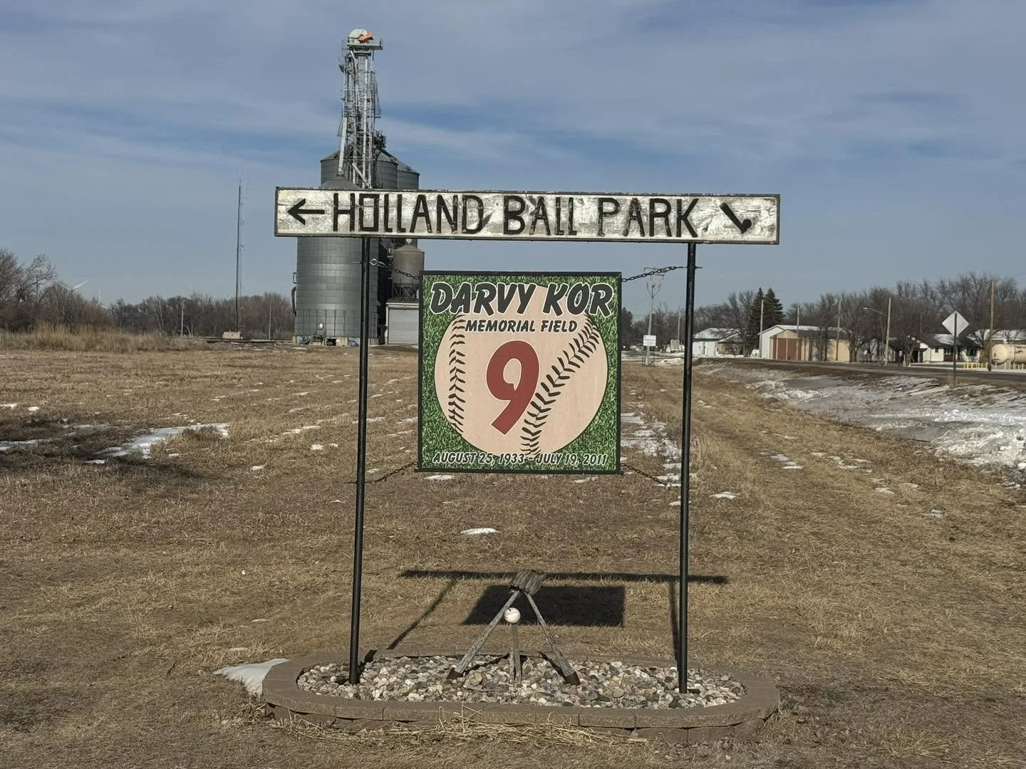 Sign featuring a large baseball with the name Darvy Kor and the number 9, in memorial for someone born August 25, 1933, and died July 19, 2011, located near a baseball field at Holland Ball Park.