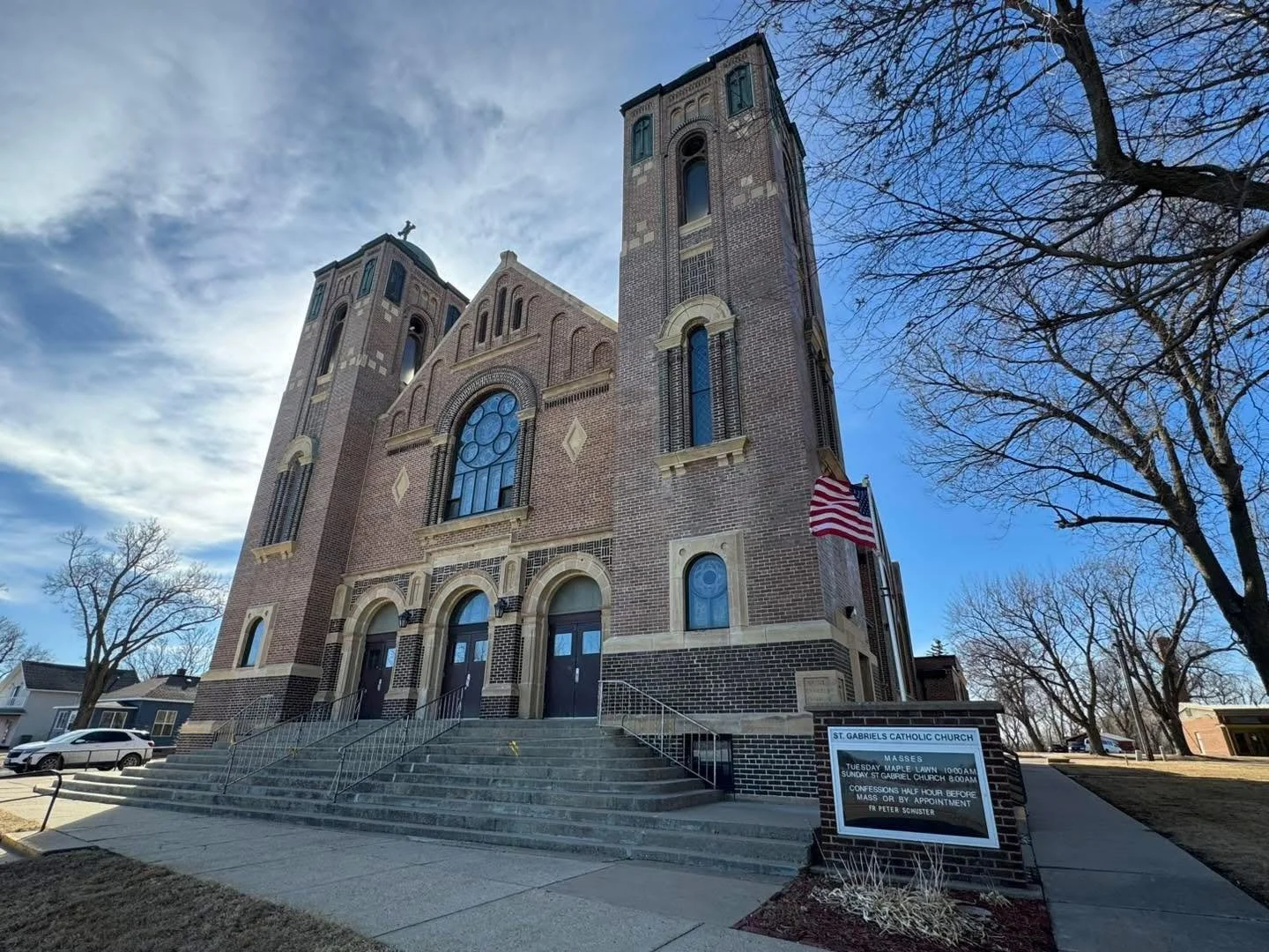 Front view of St. Gabriel's Catholic Church, a large brick church with two tall towers and stained glass windows, with a ramp and stairs leading to the entrance, and an American flag outside.