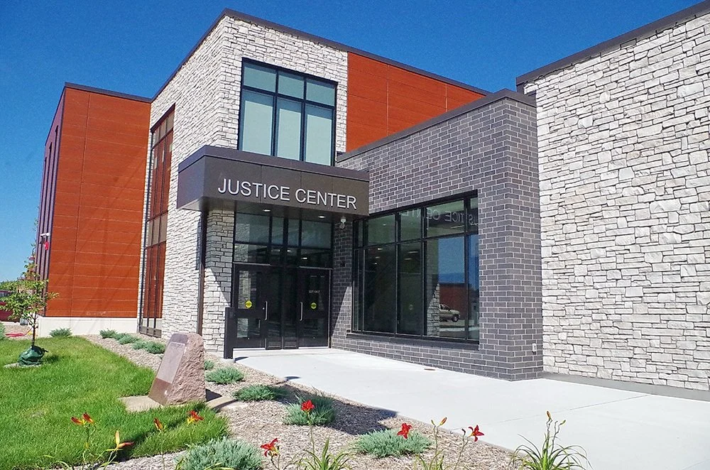 Front entrance of a modern justice center building with a sign, glass doors, and mixed brick and stone exterior walls, surrounded by greenery and flowers.