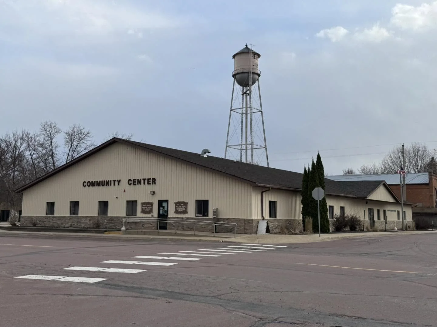 A small community center building with a water tower in the background, situated along a street with a crosswalk in front.