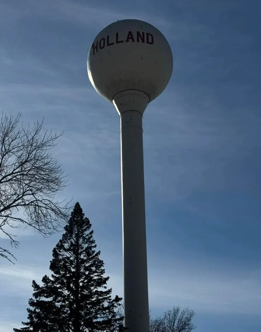 A tall white water tower with the word 'HOLLAND' written on the spherical top, set against a clear blue sky with some tree branches in the foreground.