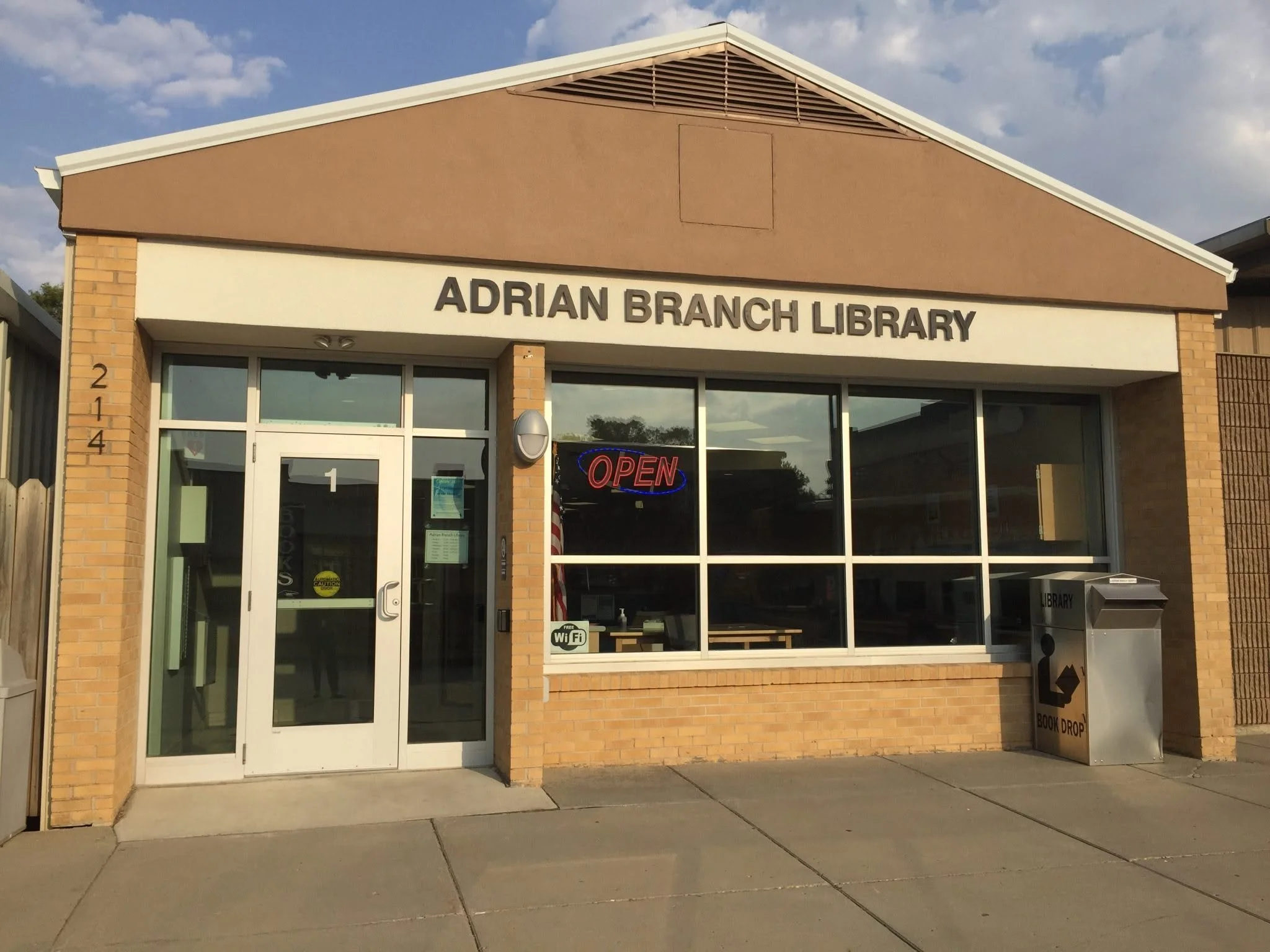 Front view of Adrian Branch Library building with large windows, sign, and an open sign in the window, next to a book drop container.