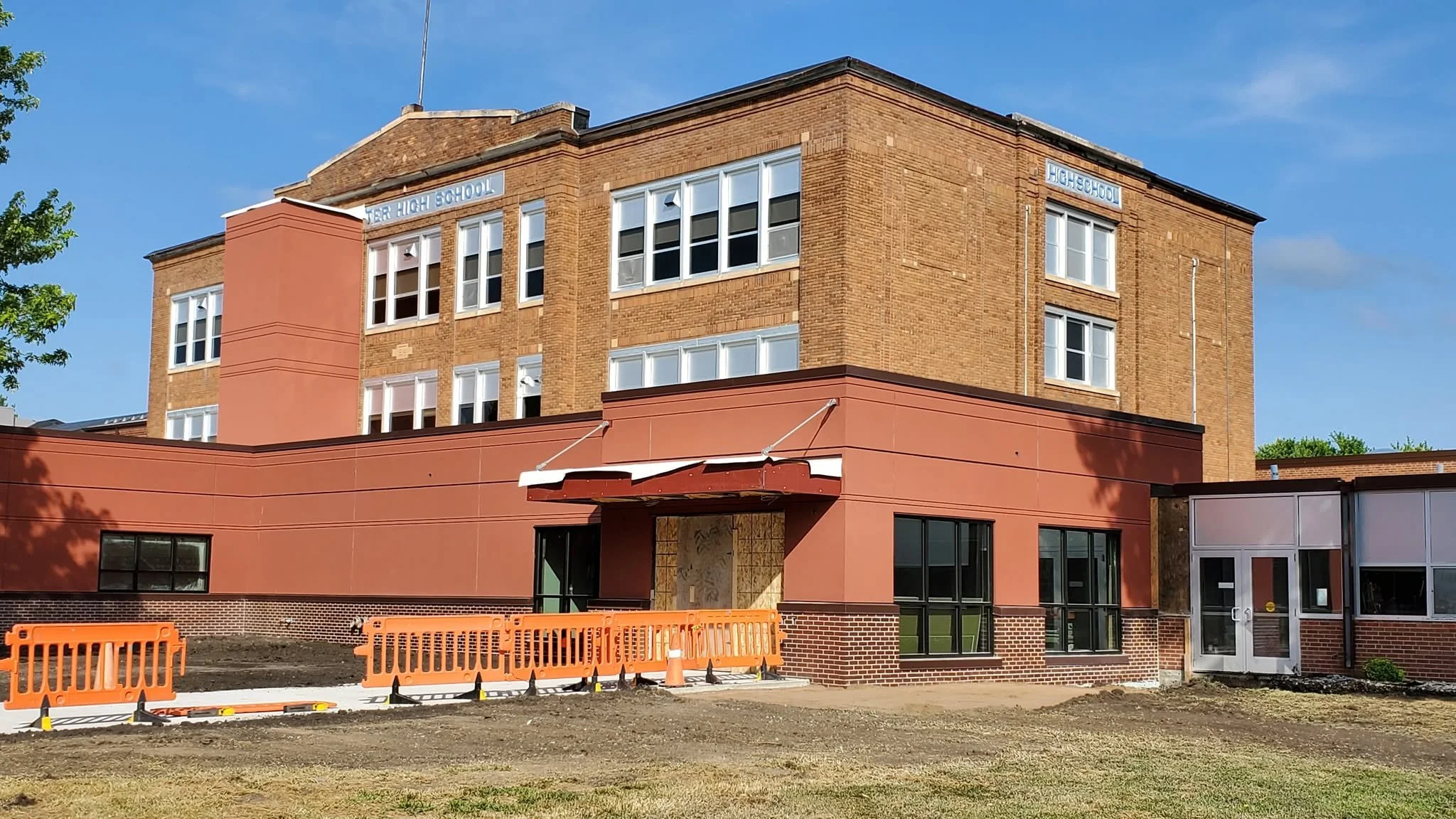 Construction site of a modern brick school building with an orange facade and large windows, orange safety barriers, and a clear blue sky.