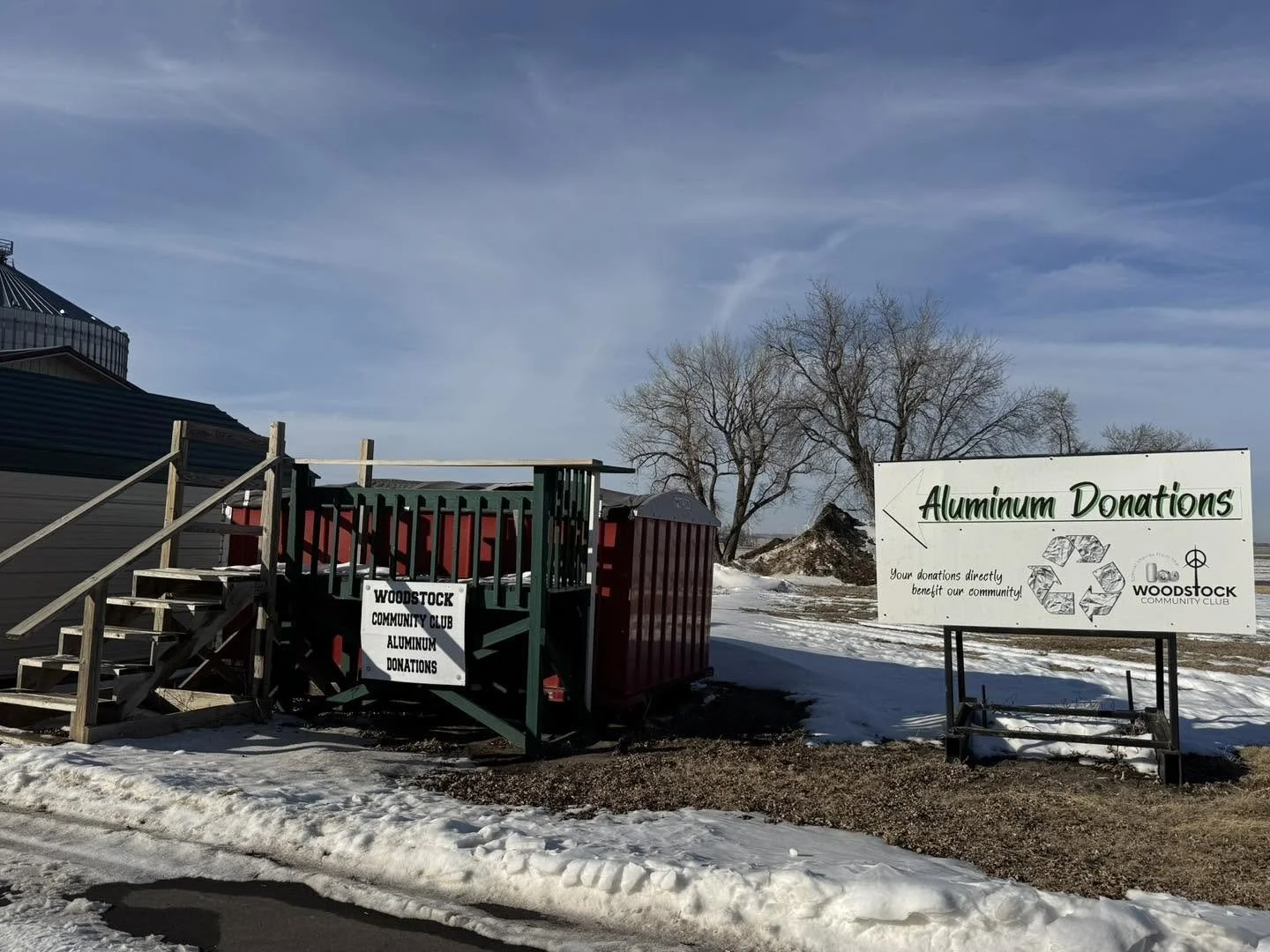 Outdoor scene with a sign that reads 'Aluminum Donations' and a wooden staircase leading to a red container for donations from Woodstock Community Club. Surrounding area has snow and leafless trees, indicating winter.