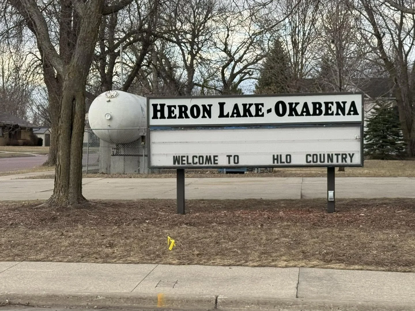 Sign reading "Heron Lake-Okabena" and "Welcome to HLO Country" in a small town setting with bare trees and houses in the background.