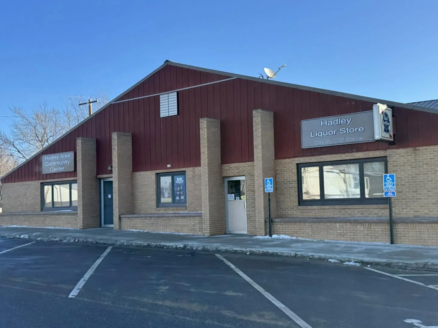 A brick building with a red metal roof houses the Hadley Area Community Center and Hadley Liquor Store, which is on and off sale. There are two parking spaces and two accessible parking signs in front of the building, with some snow on the pavement. The sky is clear and blue.
