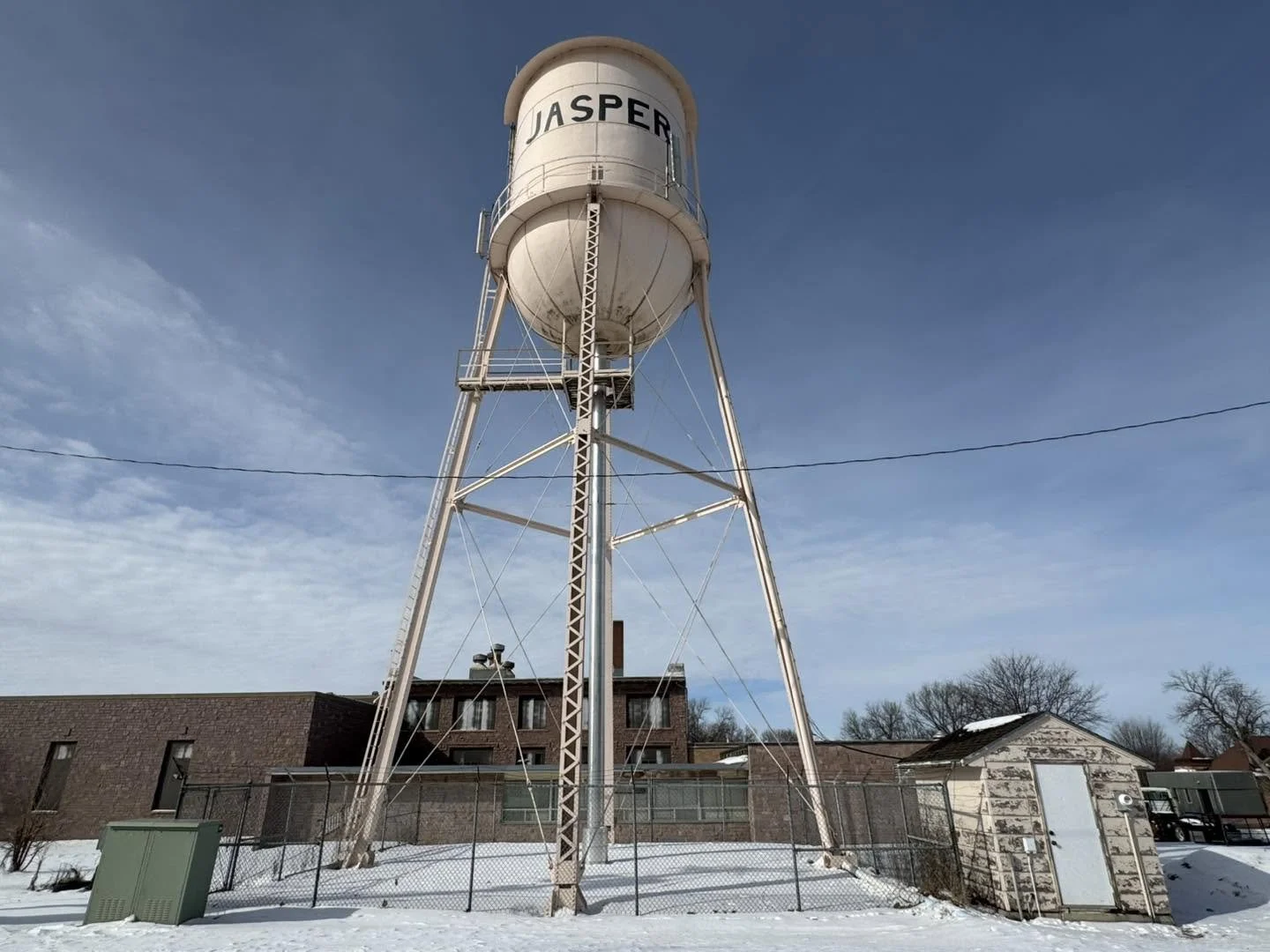 A tall white water tower with the name 'JASPER' painted on it, surrounded by snowy ground, fences, and nearby buildings under a blue sky.
