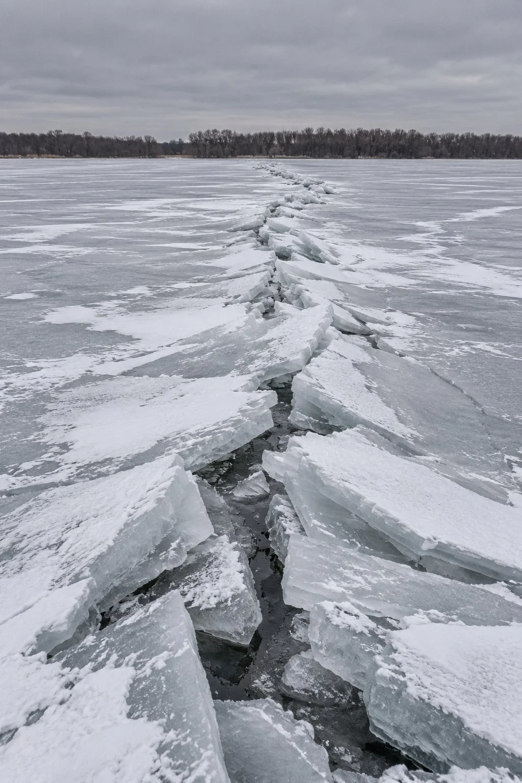 Frozen lake with a large crack running through the ice, and snow-covered overcast sky with distant trees.
