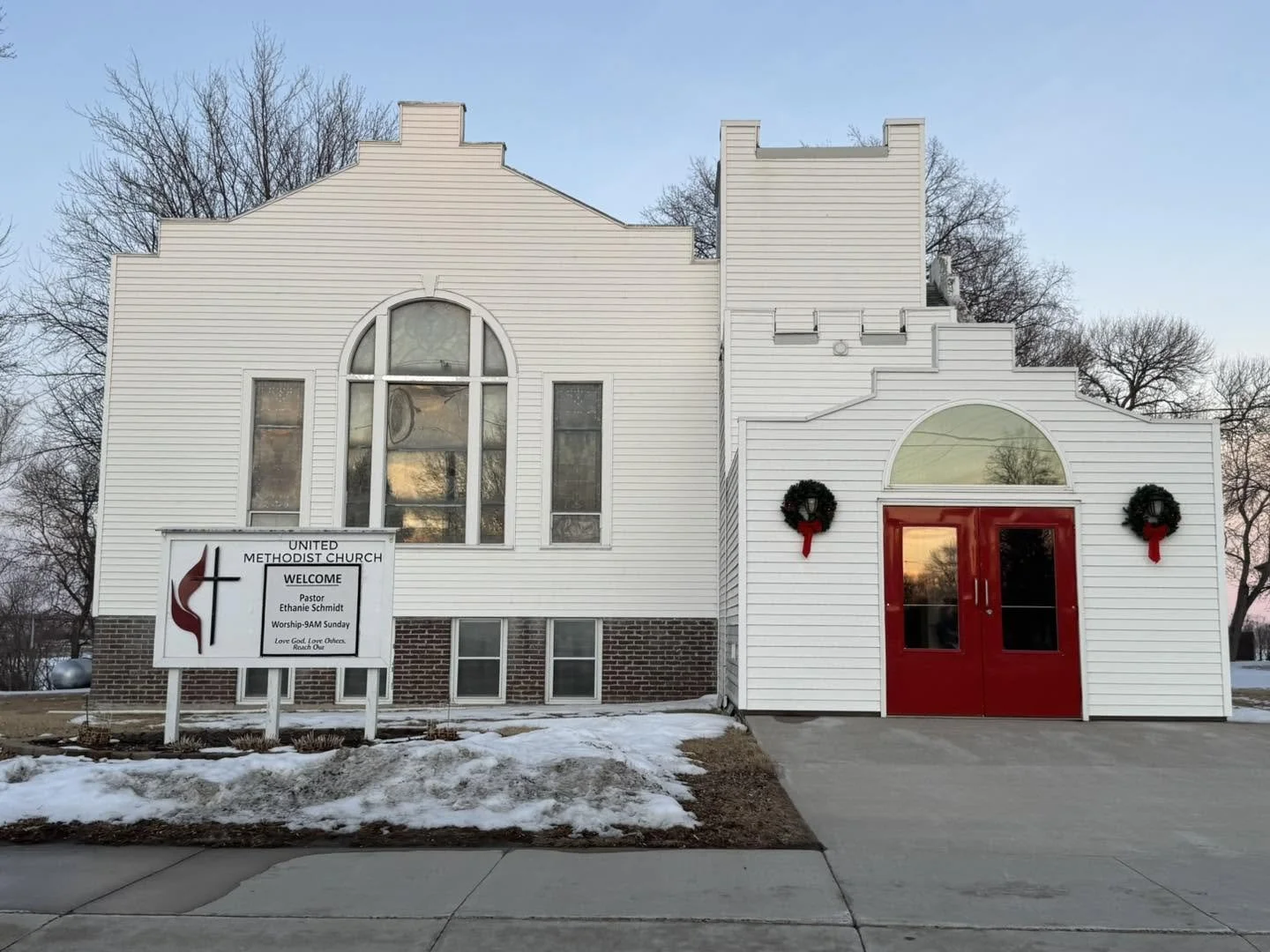 Front view of a white church building with red double doors, Christmas wreaths, and a sign indicating it's United Methodist Church, with snow on the ground.