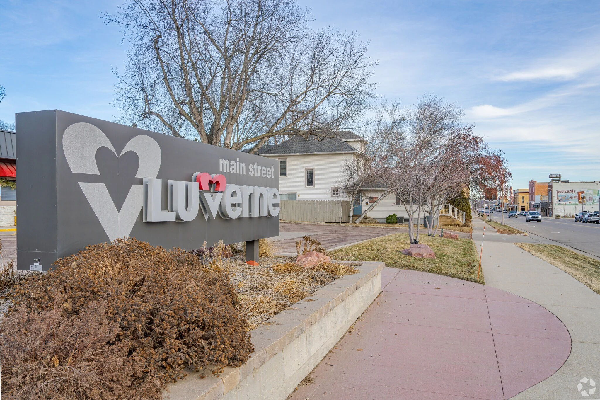 A sign reads "main street Loveline" with a stylized heart logo, located in a small town or neighborhood with a sidewalk, leafless trees, a fence, and buildings in the background under a partly cloudy sky.