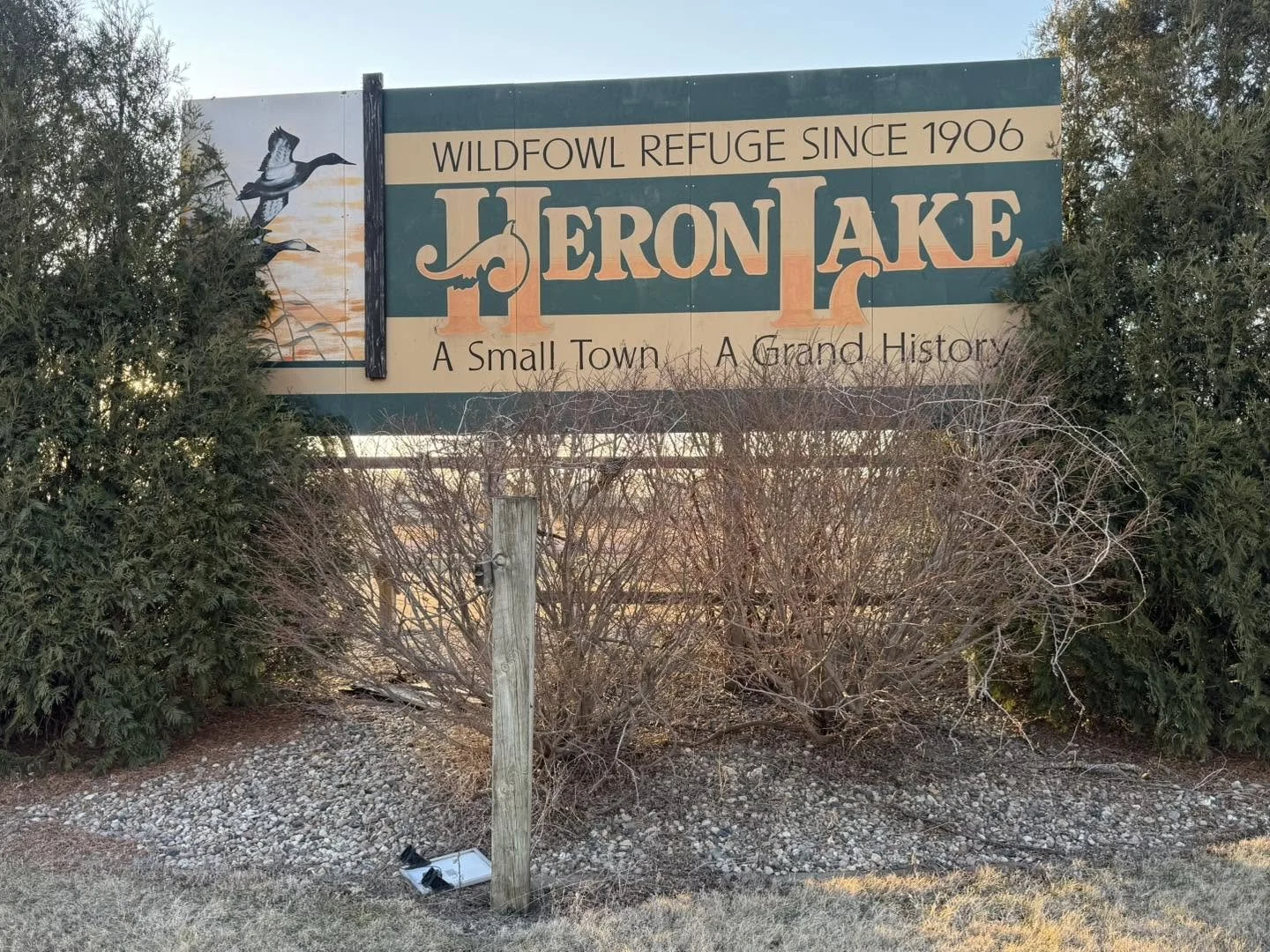 Sign for HERON LAKE, a small town and historic site, featuring a bird graphic and surrounded by bushes and gravel ground.