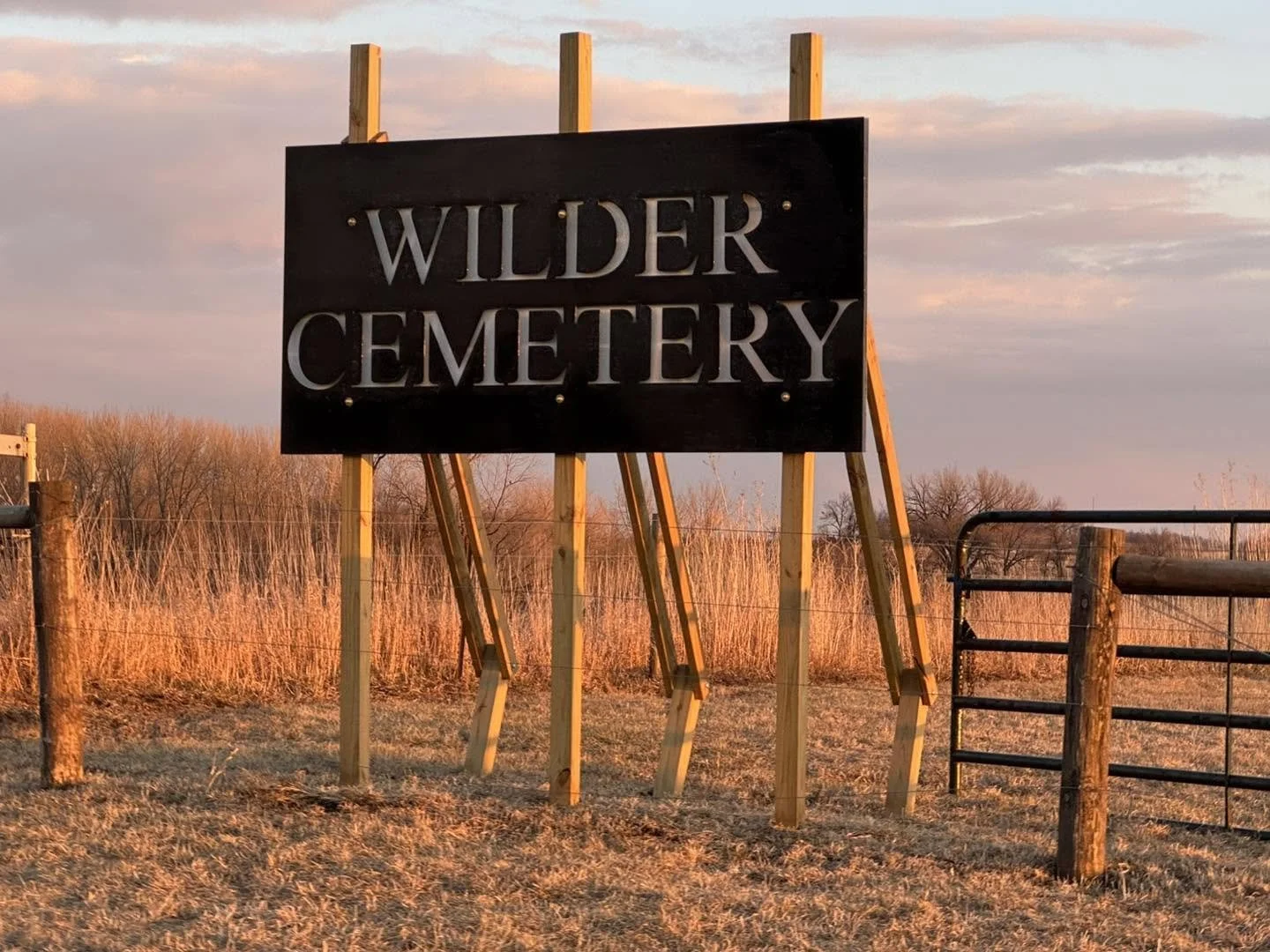A sign reads 'Wilder Cemetery' on a farm field during sunset with tall grass, a dirt path, and a wooden fence.
